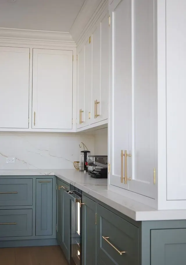 White upper cabinets with crown moulding over sage-green Shaker lowers, brass pulls and a white quartz counter