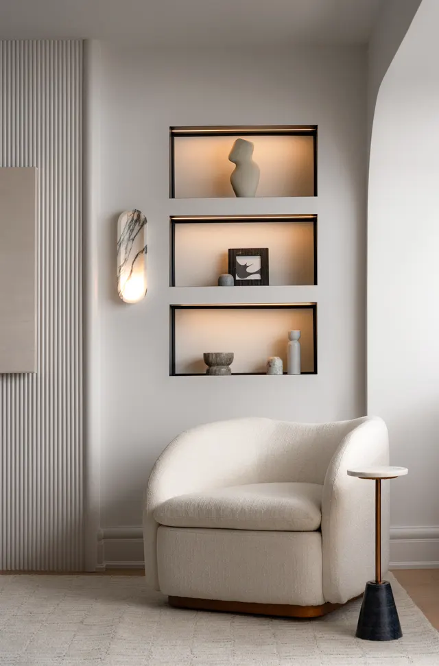 Three lit wall niches holding sculptural pieces above a cream bouclé armchair, beside a fluted panel and a marble wall sconce