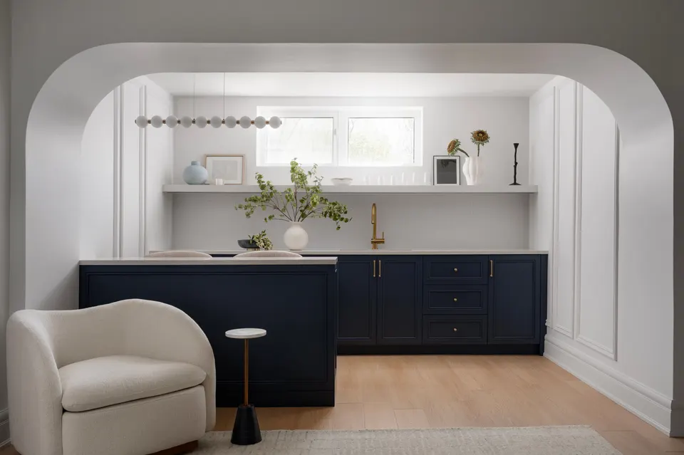Arched wet bar with navy shaker cabinets, a brass faucet and open shelving, beside a cream swivel chair