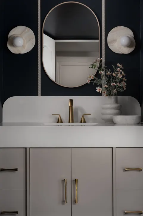 Powder room with a pill-shaped mirror between two disc sconces on a navy wall, over a cream vanity with brass pulls