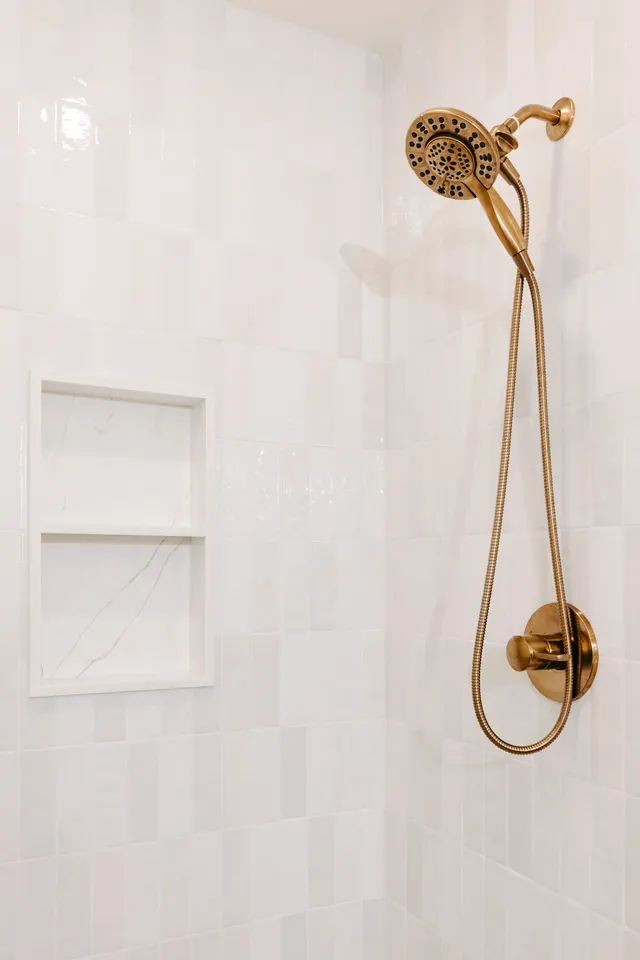 Brushed-brass hand shower on white wall tile beside a recessed niche