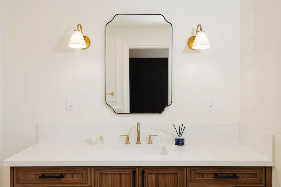 Ensuite vanity with a walnut Shaker cabinet, a white quartz top, a brushed brass faucet, a scalloped mirror and two brass wall sconces