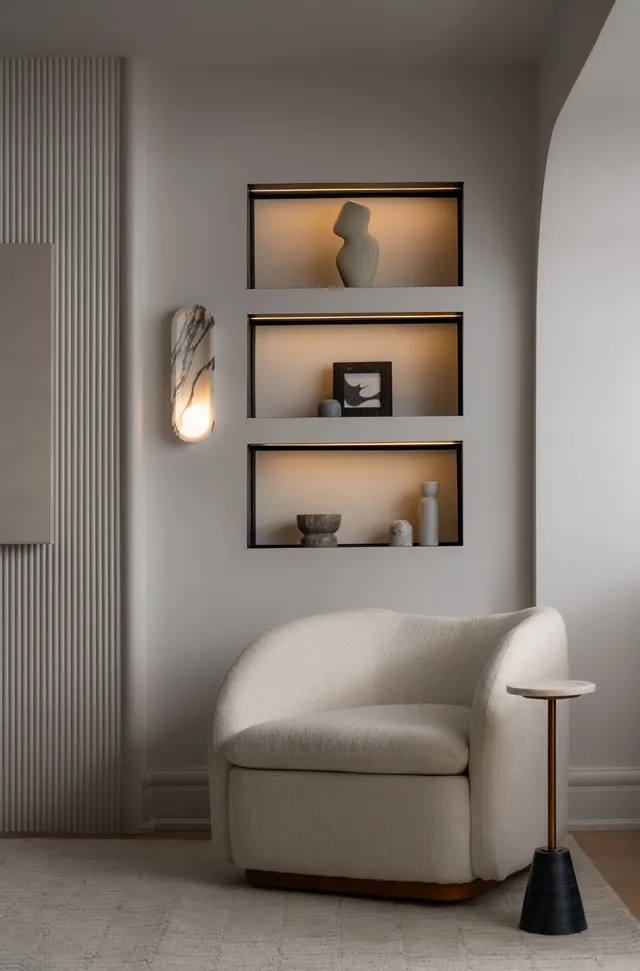 Lit display niches above a cream bouclé chair and a marble wall sconce