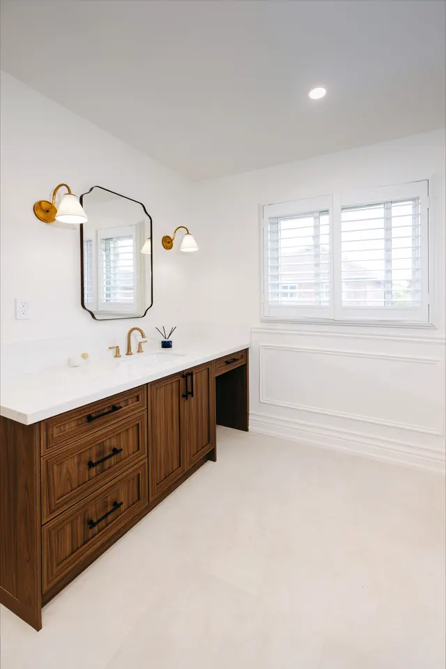 Walnut vanity with brass sconces and a scalloped mirror under shuttered windows
