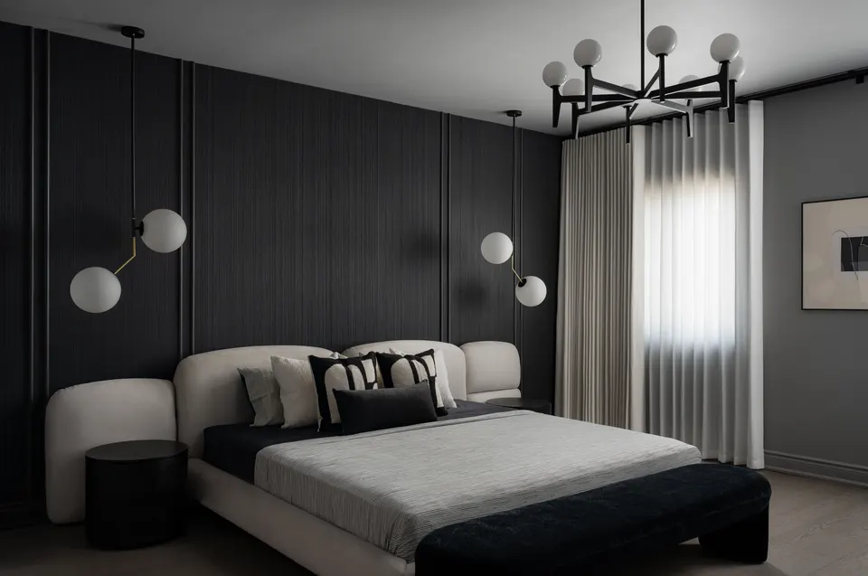 Bedroom with a fluted black feature wall, a curved cream upholstered bed, two-globe pendants, a black globe chandelier and sheer curtains at the window