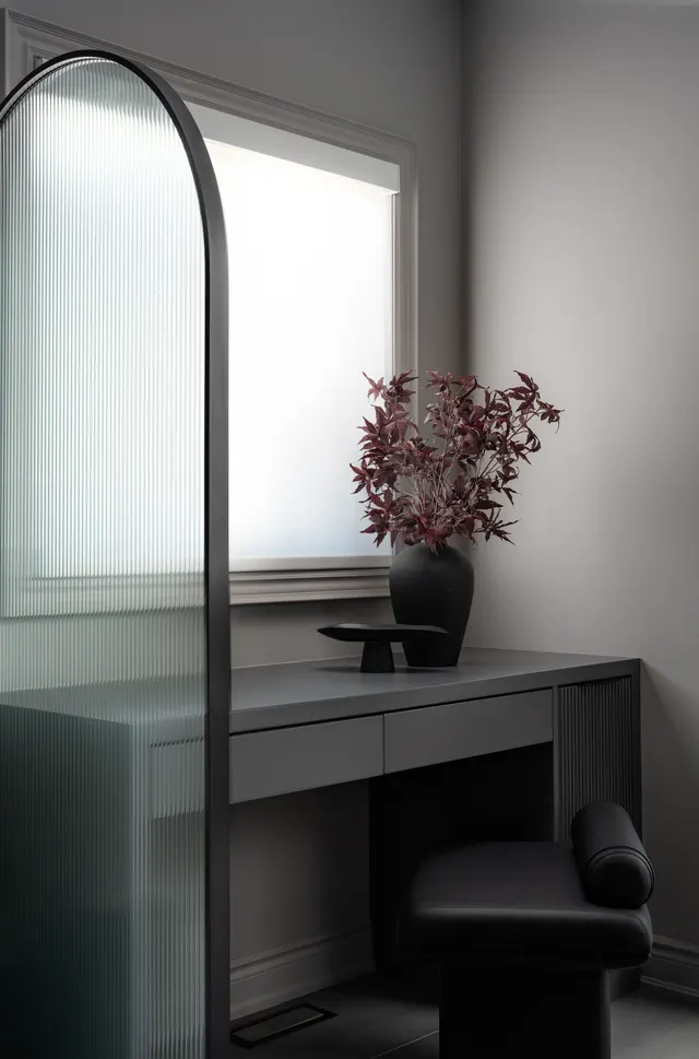 Vanity nook with an arched fluted-glass screen, a grey desk with drawers, a black bolster stool and a black vase of red maple leaves below a frosted window