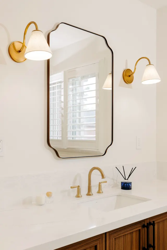 Black-framed scalloped mirror between two brass sconces over a brushed brass faucet and white quartz