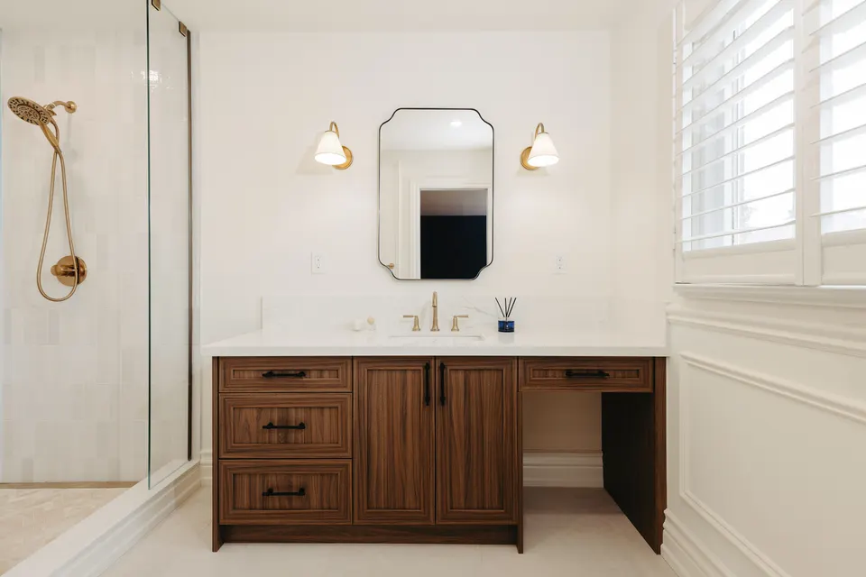 Ensuite with a frameless glass shower and brass hand shower beside a walnut vanity and makeup counter under a scalloped mirror