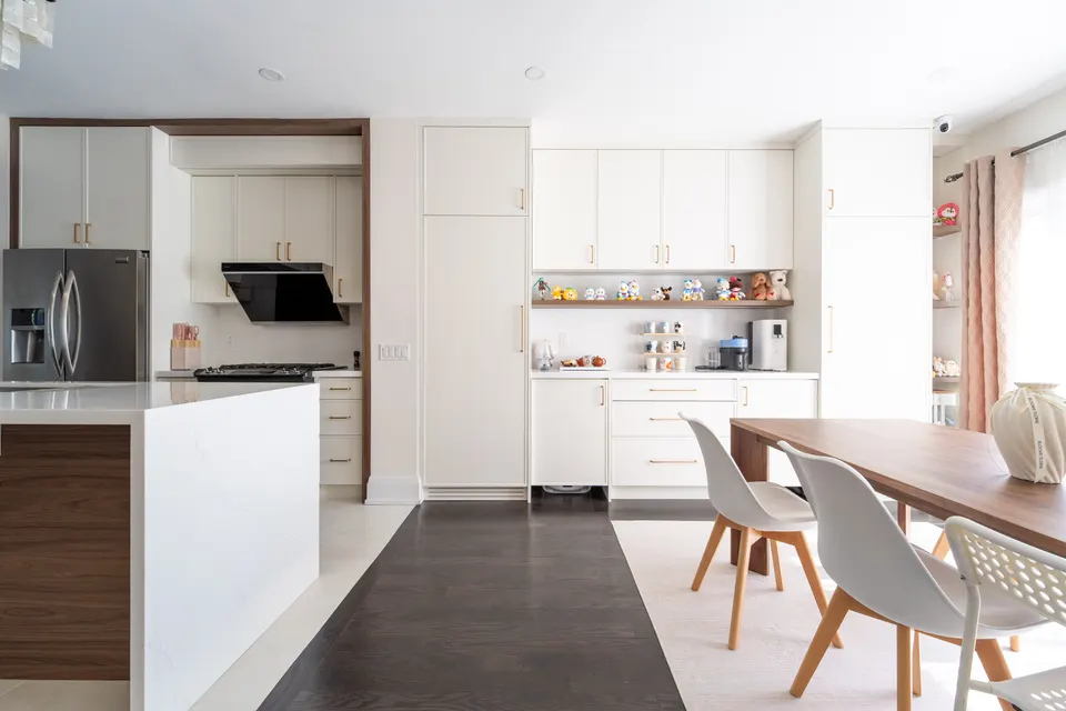 White kitchen with a walnut-panelled island opening onto a walnut dining table and white chairs on a pale rug