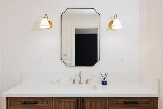 Walnut vanity with a white quartz top, a brushed-brass faucet, a black-framed mirror and brass sconces