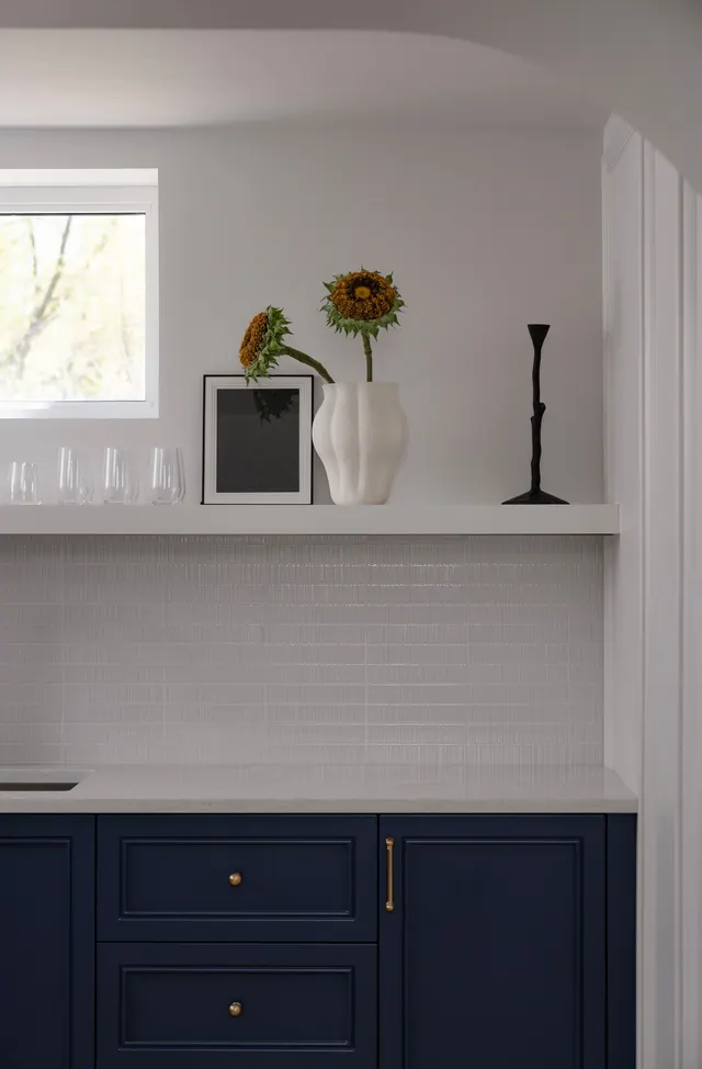 Open shelf with a sunflower in a vase and a framed print above navy cabinets and white tile