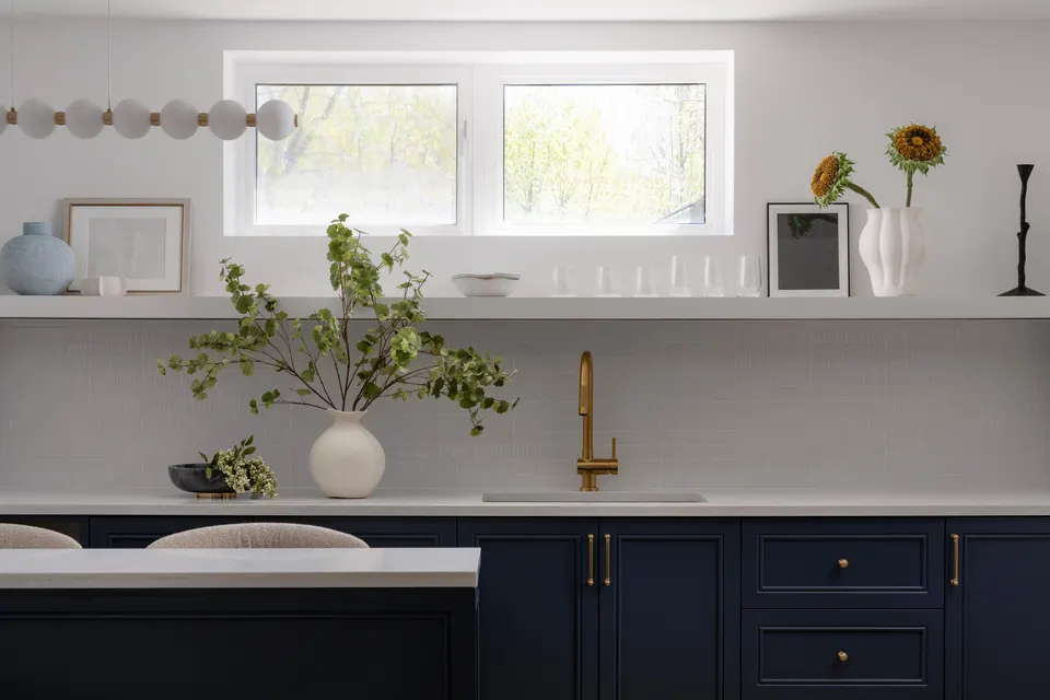 Basement wet bar with navy shaker cabinets, a white quartz counter, a brass faucet and an open shelf below the window
