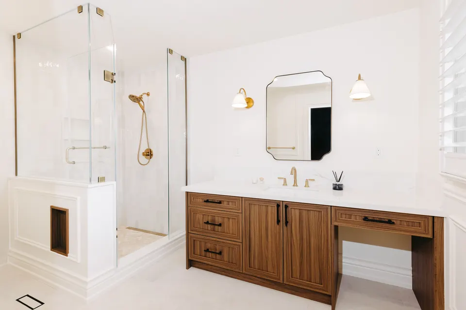 Ensuite with a frameless glass shower and brass hand shower beside a walnut vanity with a makeup counter, a scalloped mirror and a brass sconce
