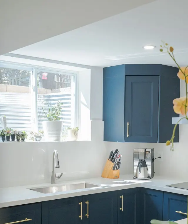 Navy shaker corner cabinet with brass pulls over a white quartz counter beside the suite’s kitchen window