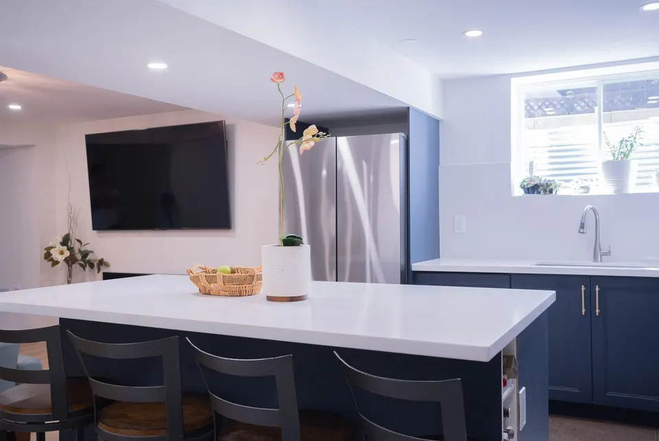 Basement suite kitchen with a white quartz island and bar stools, navy shaker cabinets, a wall-mounted TV and a window over the sink