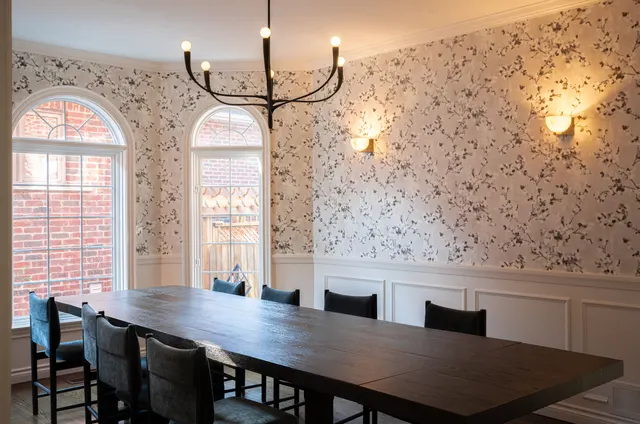 Dining room with patterned wallpaper above white wainscoting, arched windows, brass sconces and a long dark table