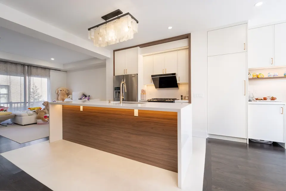 Open kitchen with a walnut-panelled island lit by an LED toe line, white quartz waterfall ends, a capiz shell chandelier and white full-height cabinetry
