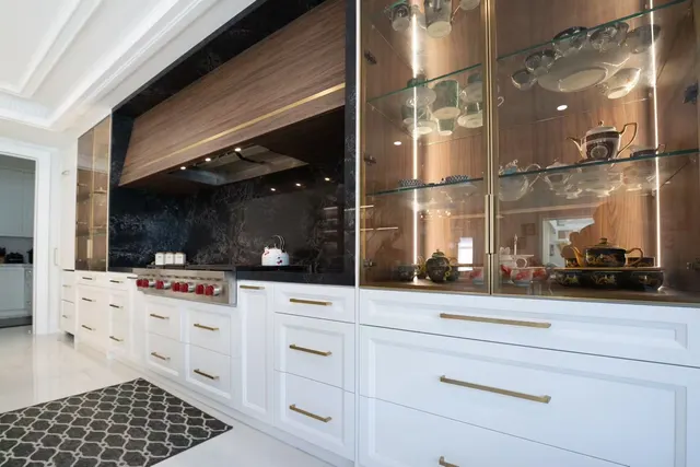 Lit glass-front display cabinets beside a walnut range hood, over white Shaker drawers with brass pulls