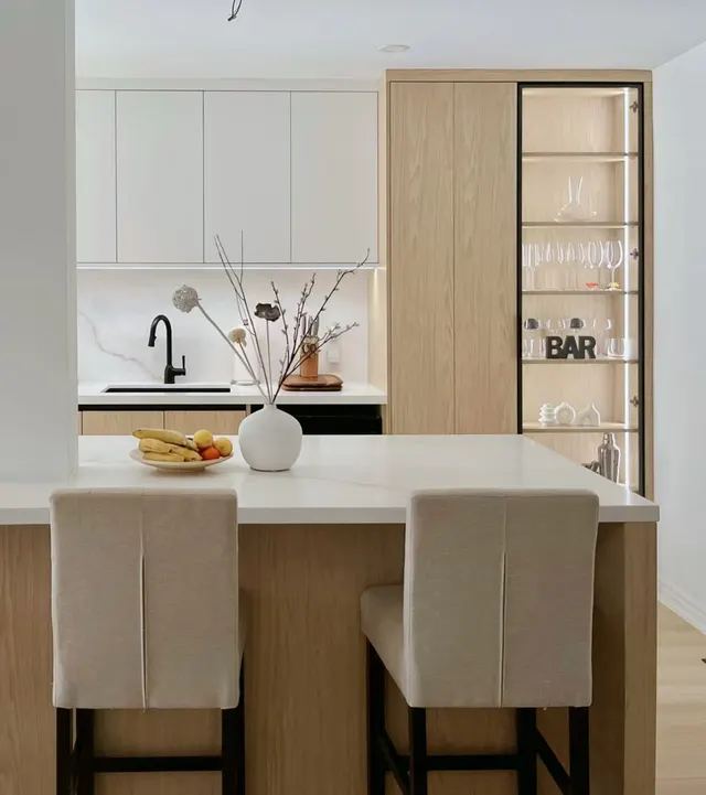Oak bar cabinet with lit glass shelves behind a white island and two linen stools