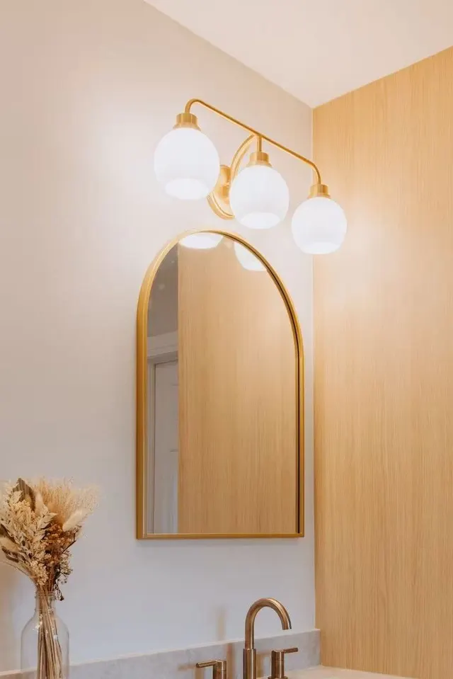Arched brass mirror and globe sconces above a vase of dried flowers and a brass faucet