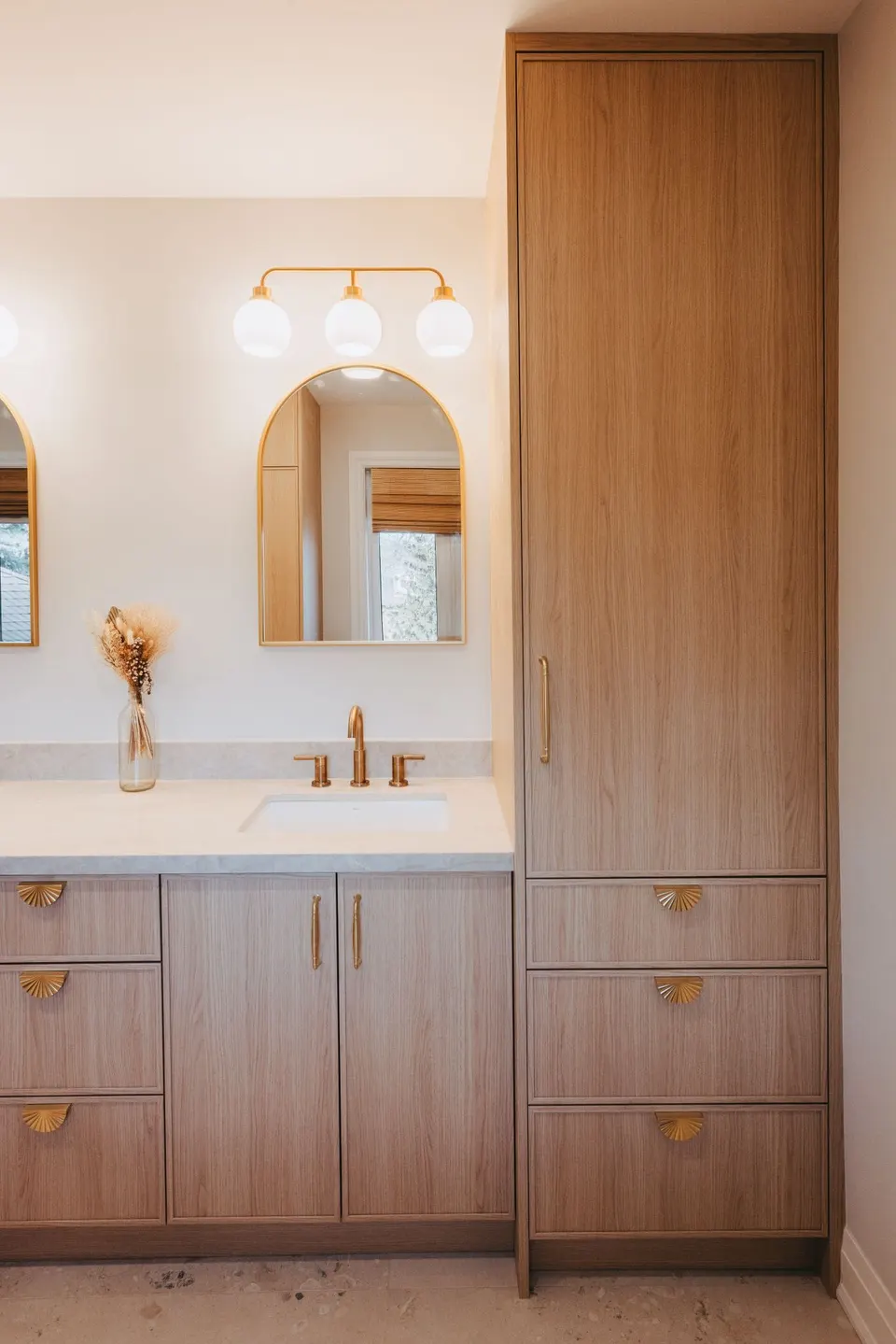 Ensuite with an oak vanity and brass faucets under an arched brass mirror and a globe sconce bar, a tall oak linen cabinet beside it