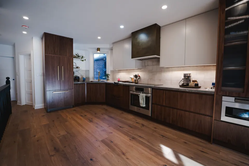 Walnut kitchen along a sunlit wall in an opened-up century home, with wide-plank oak floors and the new black stair beyond