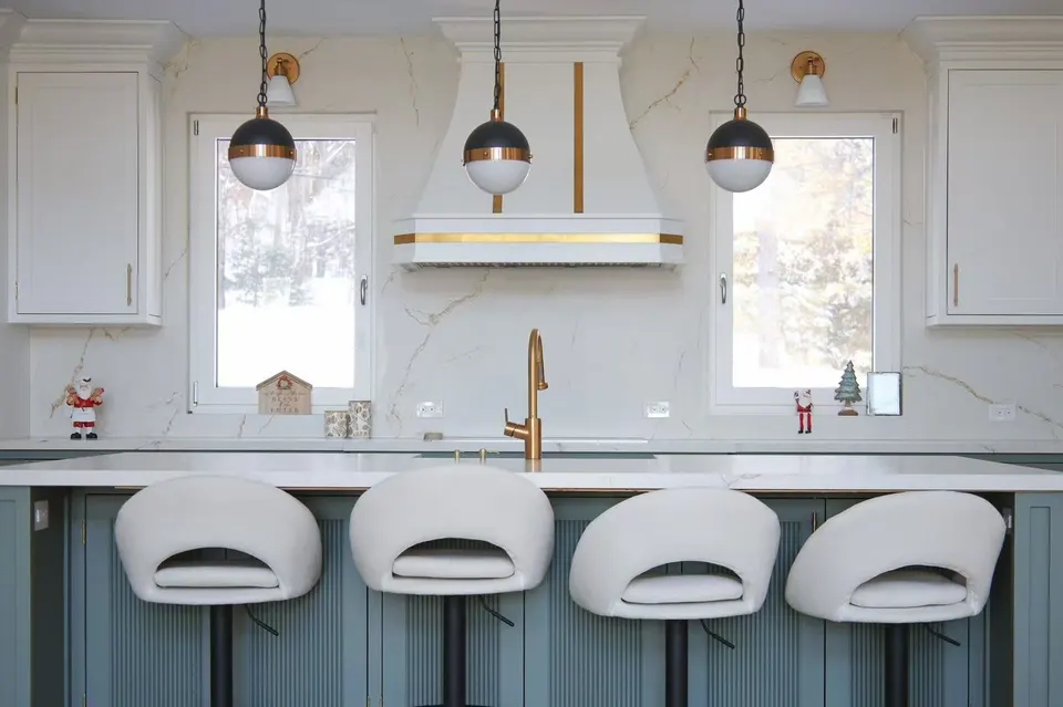 Kitchen with a sage-green island and white swivel stools under globe pendants, a white range hood banded in brass and a marble backsplash between two windows