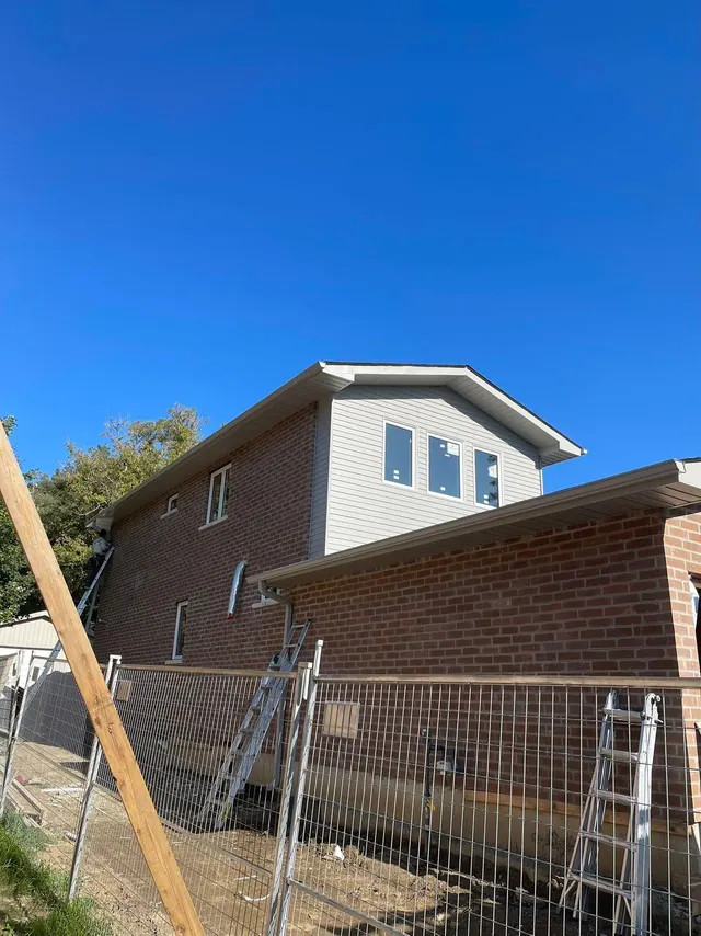 The addition’s upper storey from the side, siding and new windows above the original brick, still behind the site fence
