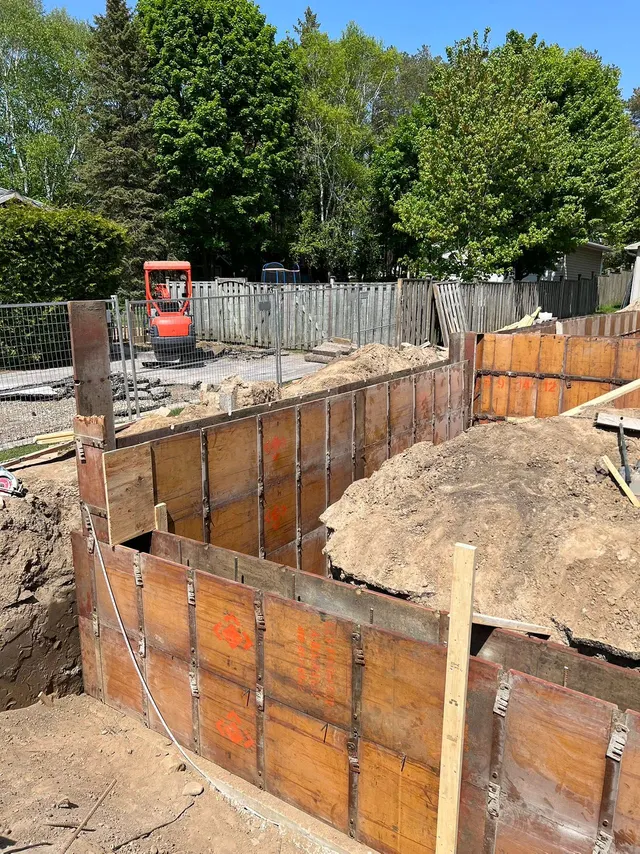 Foundation forms set in the excavation for the addition, a mini excavator behind the site fence