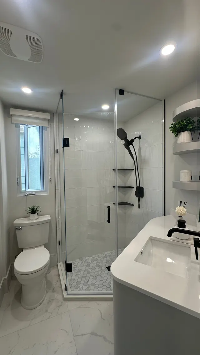 Frameless glass shower with black fixtures and a mosaic floor beside a white vanity and a small window