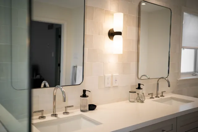 Double vanity with chrome faucets, black-framed mirrors and a frosted wall sconce against handmade tile