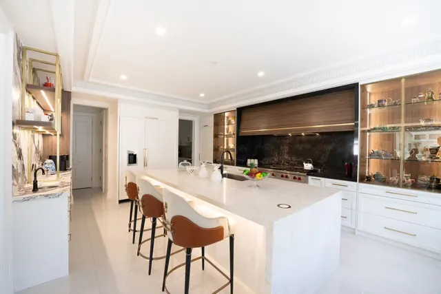 White kitchen with a lit quartz waterfall island, leather bar stools, glass-front display cabinets and a dark stone wall behind the range